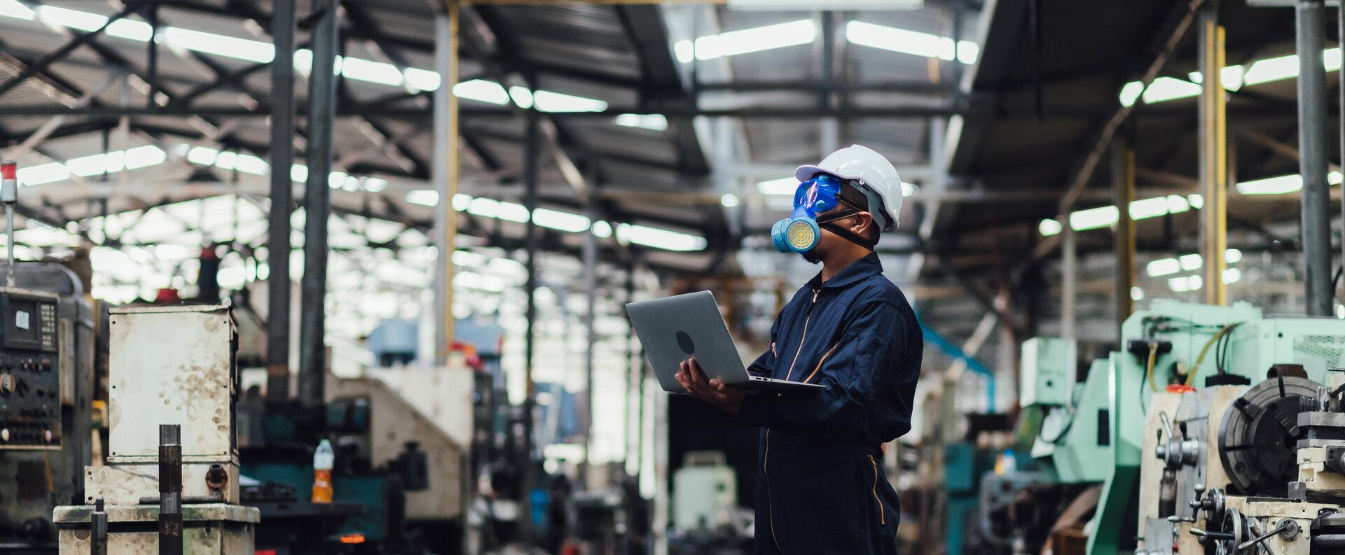 Mann mit Schutzmaske und Computerlaptop führt Kontrollarbeiten in einer Fabrik durch.