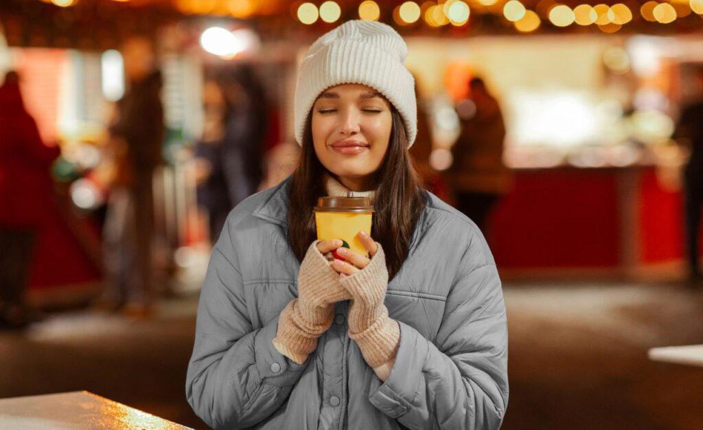 Frau haelt einen Glühweinbecher auf dem Weihnachtsmarkt in der Hand und genießt ein heißes Getränk